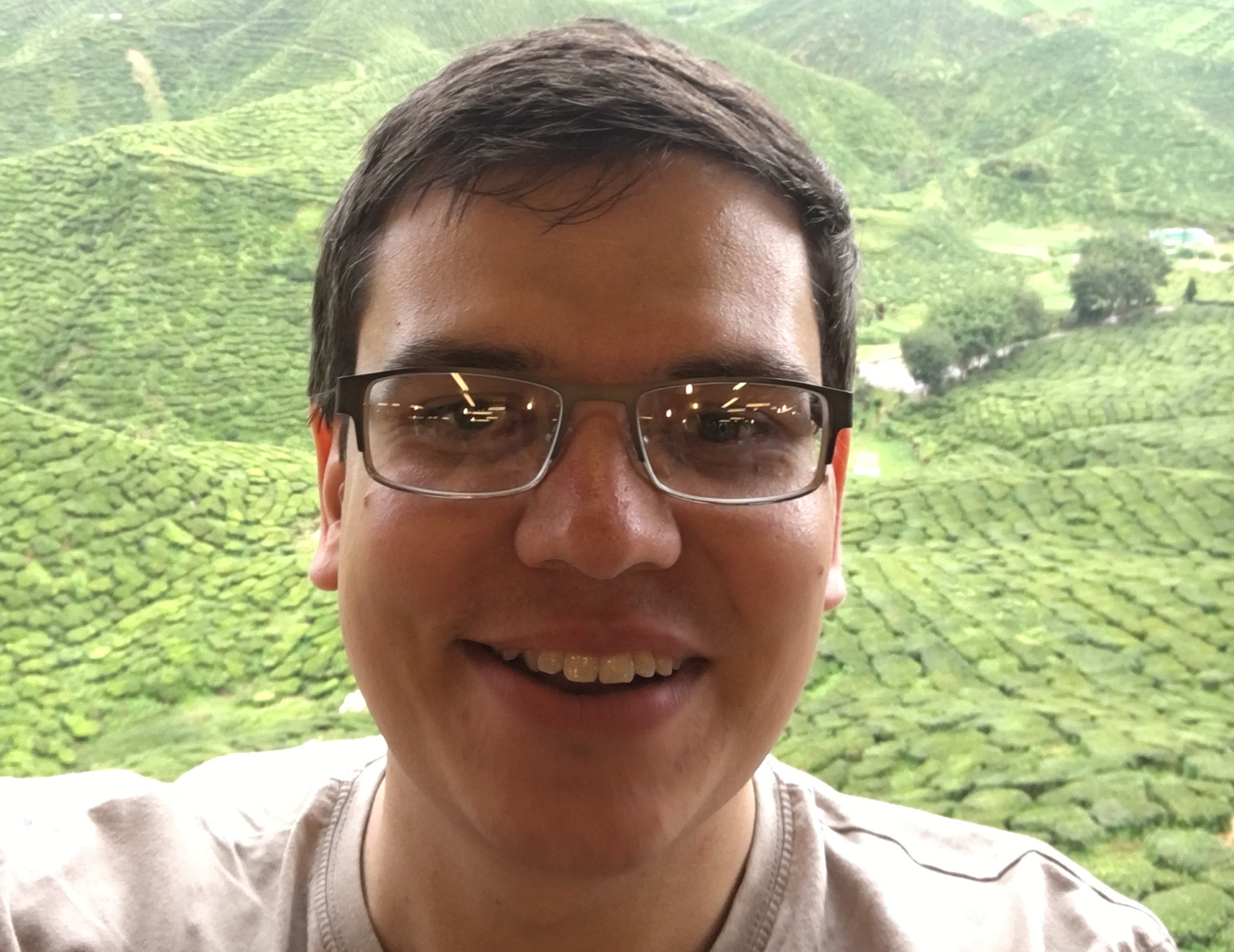 Man with brown hair and glasses, taking a selfie in front of a tea plantation